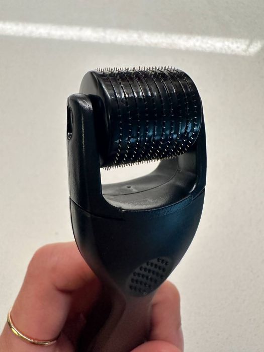 A close-up of a hand holding a derma roller, showing the roller head with numerous fine needles. 