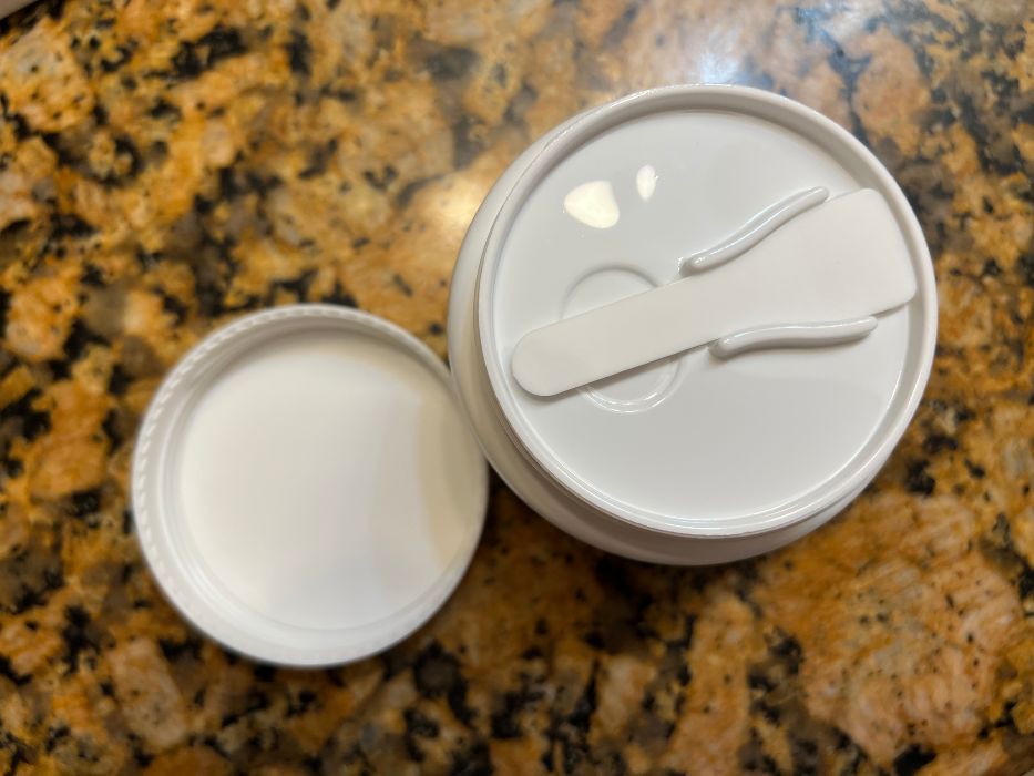 A jar of cleansing balm with the lid off, showing a spatula stored on the inner cover.
